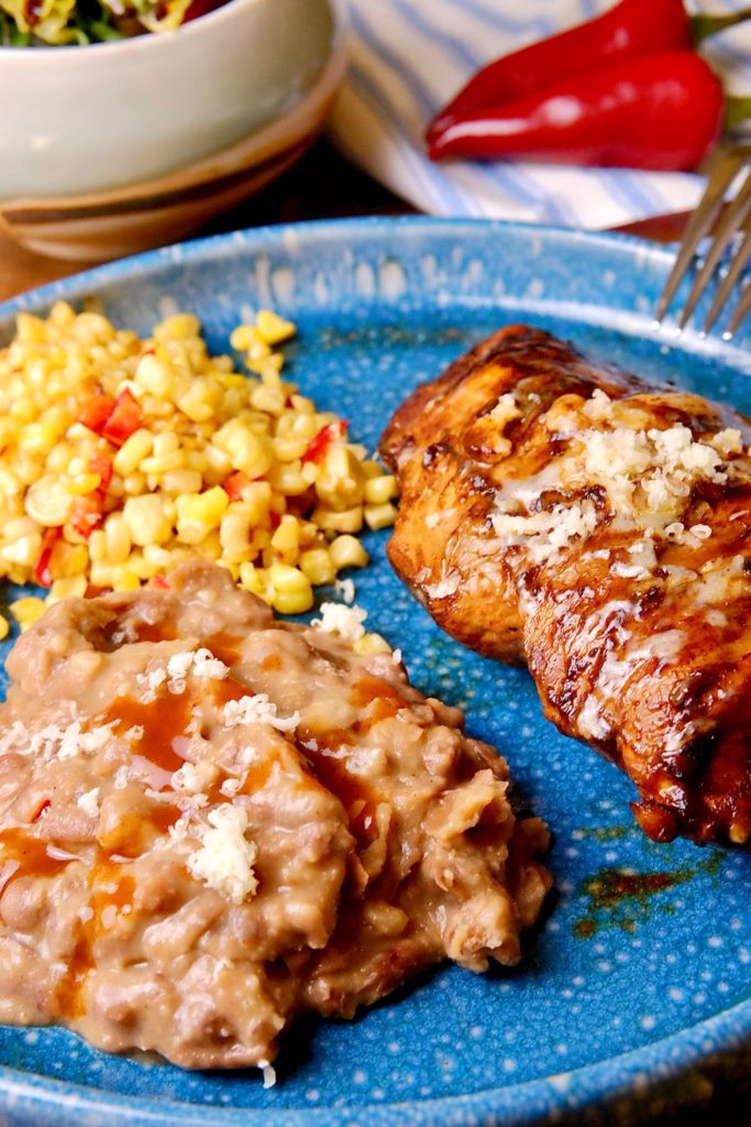 Refried beans served alongside Guajillo Chili Chicken and confetti corn on a blue stoneware plate.  Red chili peppers on blue striped linen and bowl of salad in background.