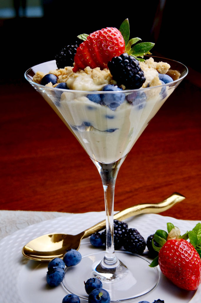 Vanilla pudding layered with blueberries in a tall martini glass set on a white plate. Pudding is garnished with cookie sand, blackberries, strawberries and mint. Additional blueberries, blackberries and strawberries are scattered around the stem of glass next to a gold spoon.