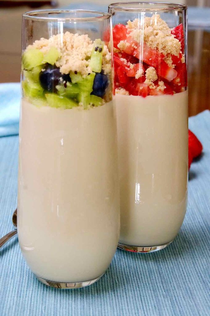 Parfait glasses filled with Vanilla Pudding.  One topped with kiwi and blueberries.  One topped with strawberries.  Both are topped with Edible Sand with light blue linen underlay.  