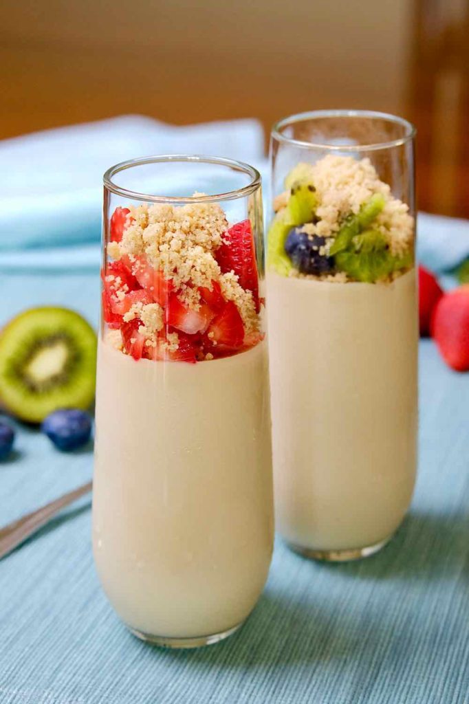 Tall parfait glasses filled with Vanilla Pudding made with Almond Milk, and garnished with diced strawberries, kiwi and blueberries set on a light blue linen with spoon and fruit in background.