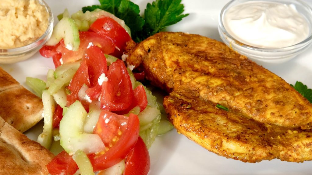 Grilled chicken breast on a white plate served with tomato cucumber salad, pita, hummus and yogurt sauce.