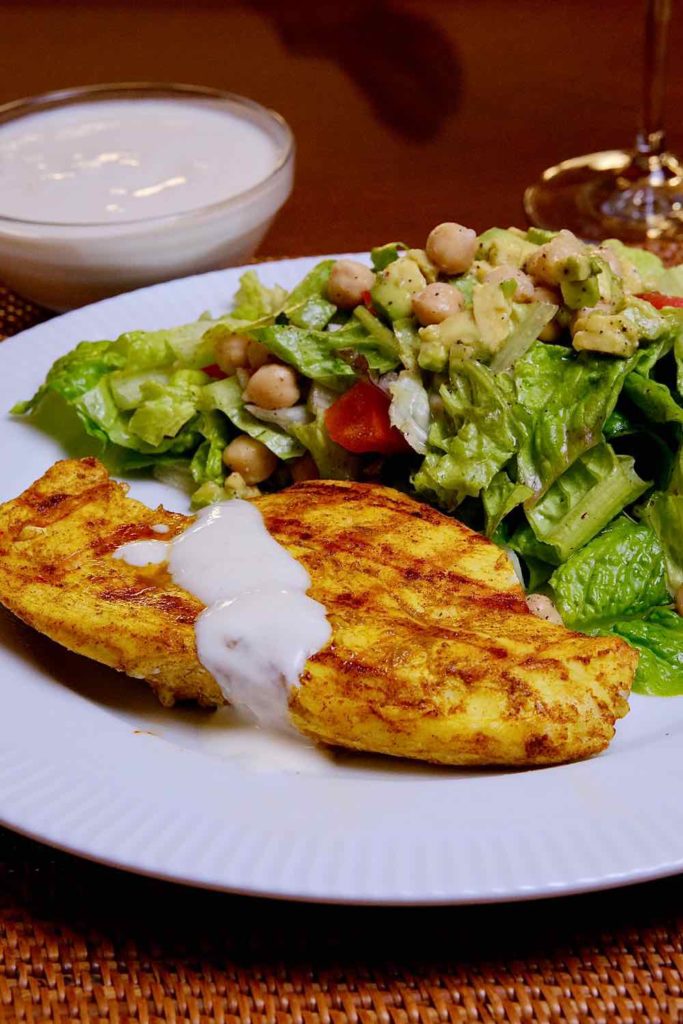 Chicken Shawarma on a white plate served with drizzled yogurt sauce drizzled and Mediterranean Salad. Bowl of yogurt sauce is set in background.