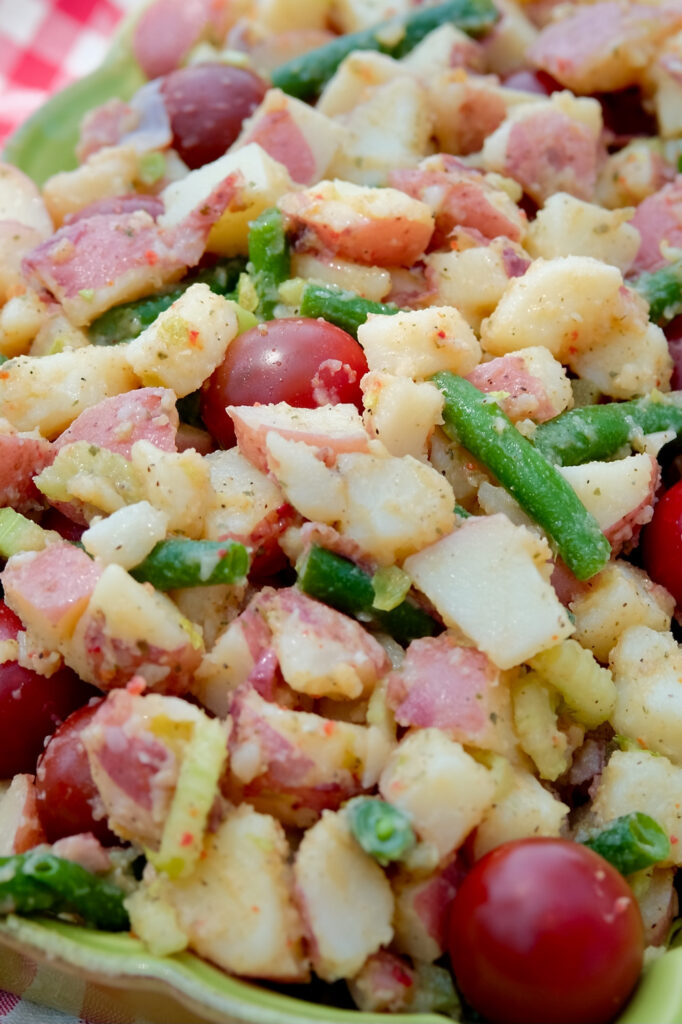 Close up shot of potato salad-cubed potatoes, green beans and cherry tomatoes dressed with an Italian herb dressing served in a rectangular ruffled edged serving dish with serving spoon resting in right forefront of dish.