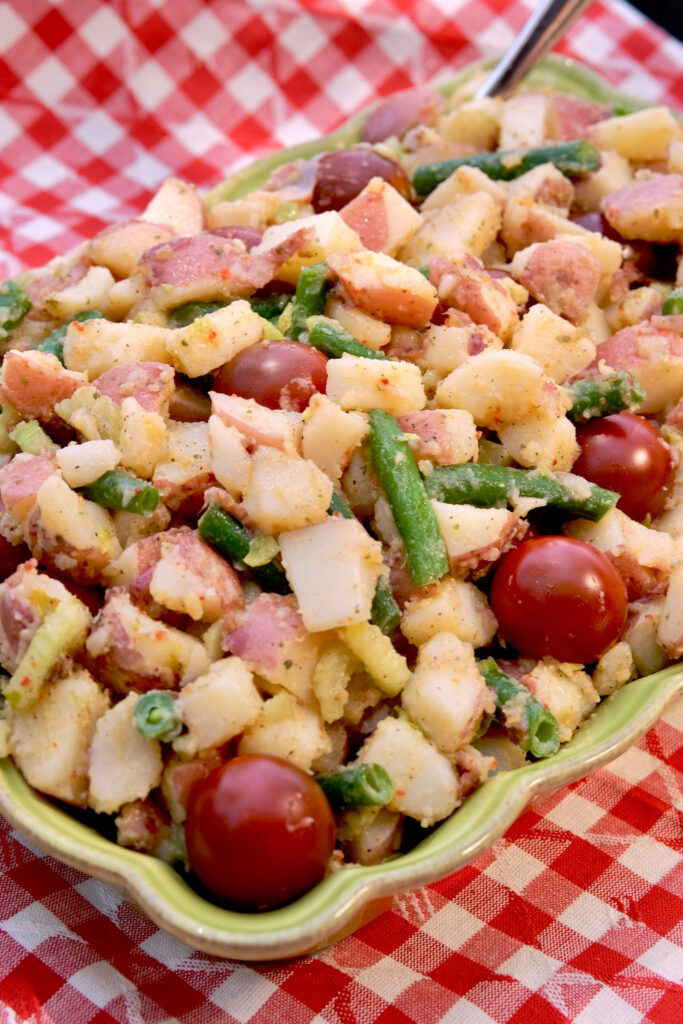 Cubed potatoes, green beans and cherry tomatoes dressed with an Italian herb dressing served in a rectangular ruffled edged serving dish is set on a red and white checkered linen.