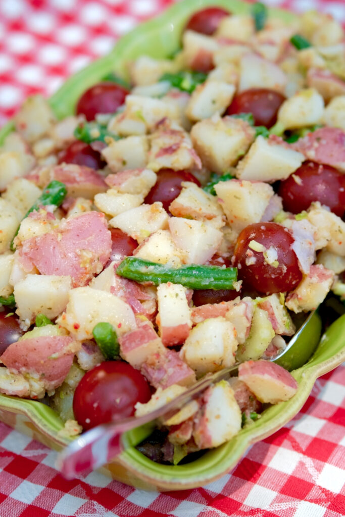 Cubed potatoes, green beans and cherry tomatoes dressed with an Italian herb dressing served in a rectangular ruffled edged serving dish is set on a red and white checkered linen.