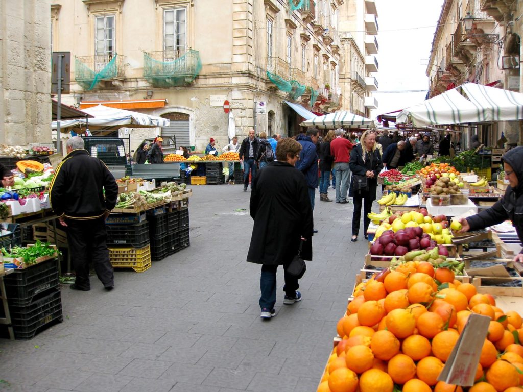 Mia looking at stands of oranges, bananas and vegetables