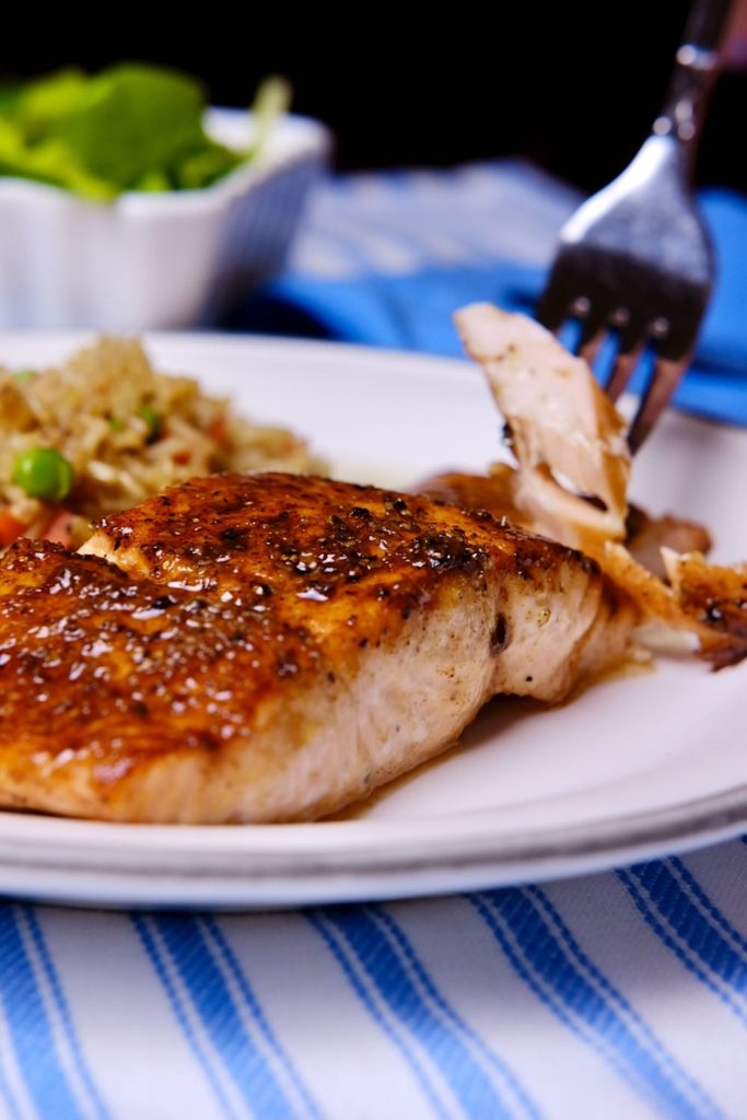 Pan Seared Barbecue Salmon and rice pilaf on white plate featuring bite shot on fork.  Salad bowl of fresh green in background set on blue and white linen.