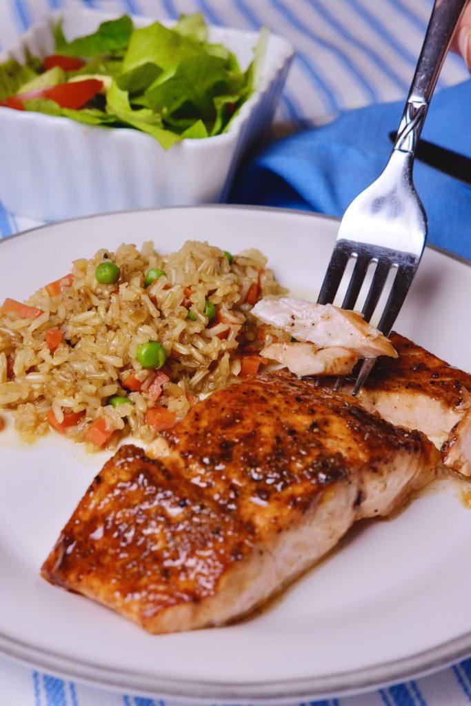 Pan Seared Barbecue Salmon on white plate with rice pilaf, showing bite shot on fork.  Bowl of salad greens in background set on blue and white linen.