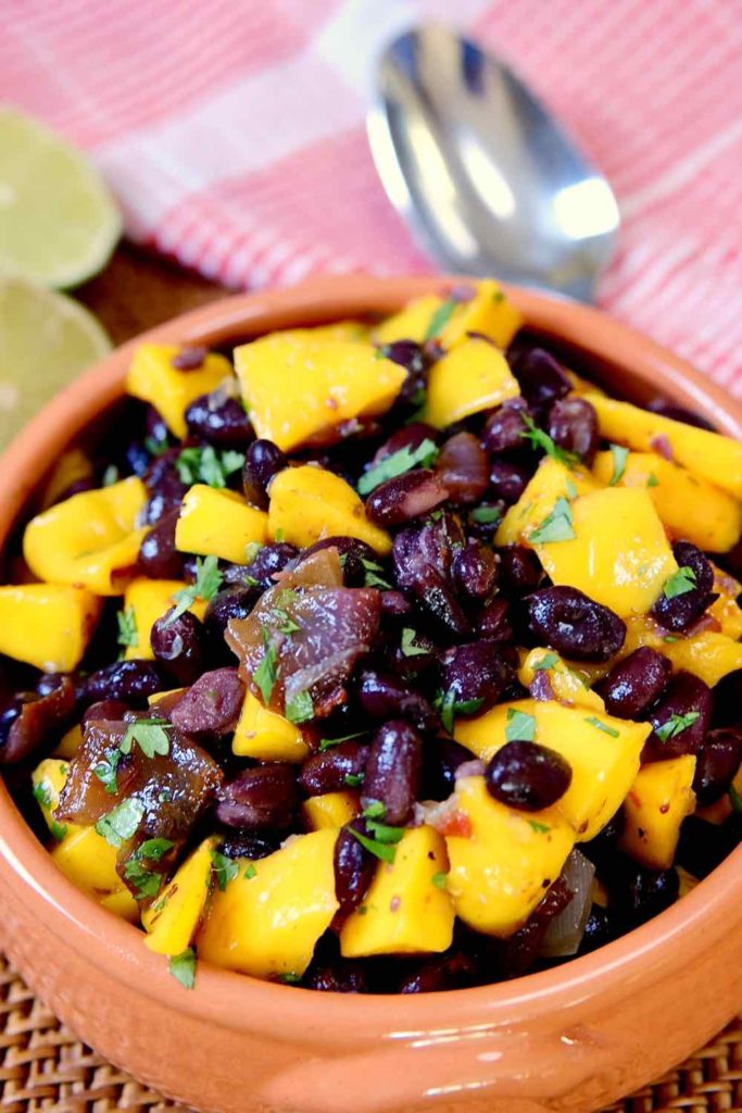 Spicy Black Bean Salad with Mangos in terracotta bowl on wicker mat with spoon on red linen and cut limes in background.