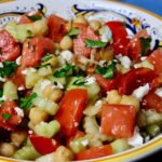 Tomato Cucumber Salad with chickpeas served in a blue and gold Italian bowl garnished with crumbled Feta cheese and fresh chopped parsley.