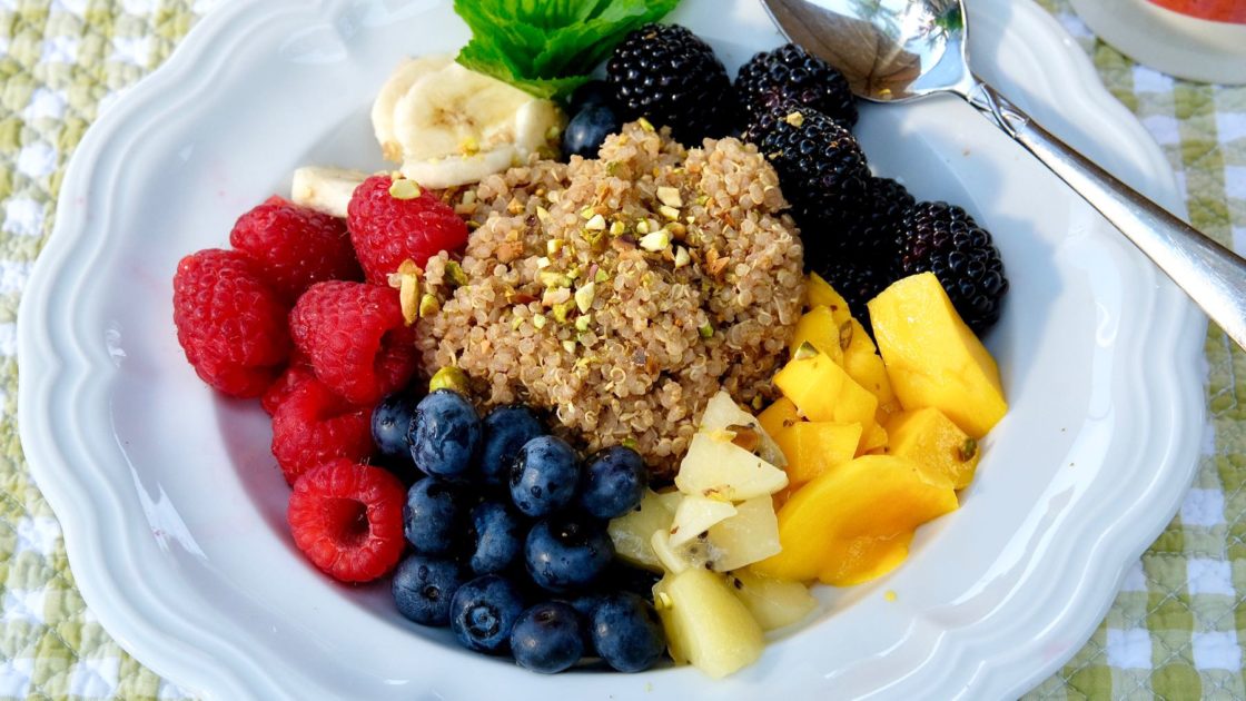 Quinoa in white bowl surrounded by blueberries, raspberries, sliced banana, blackberries and chopped mango, sprinkled with chopped pistachios