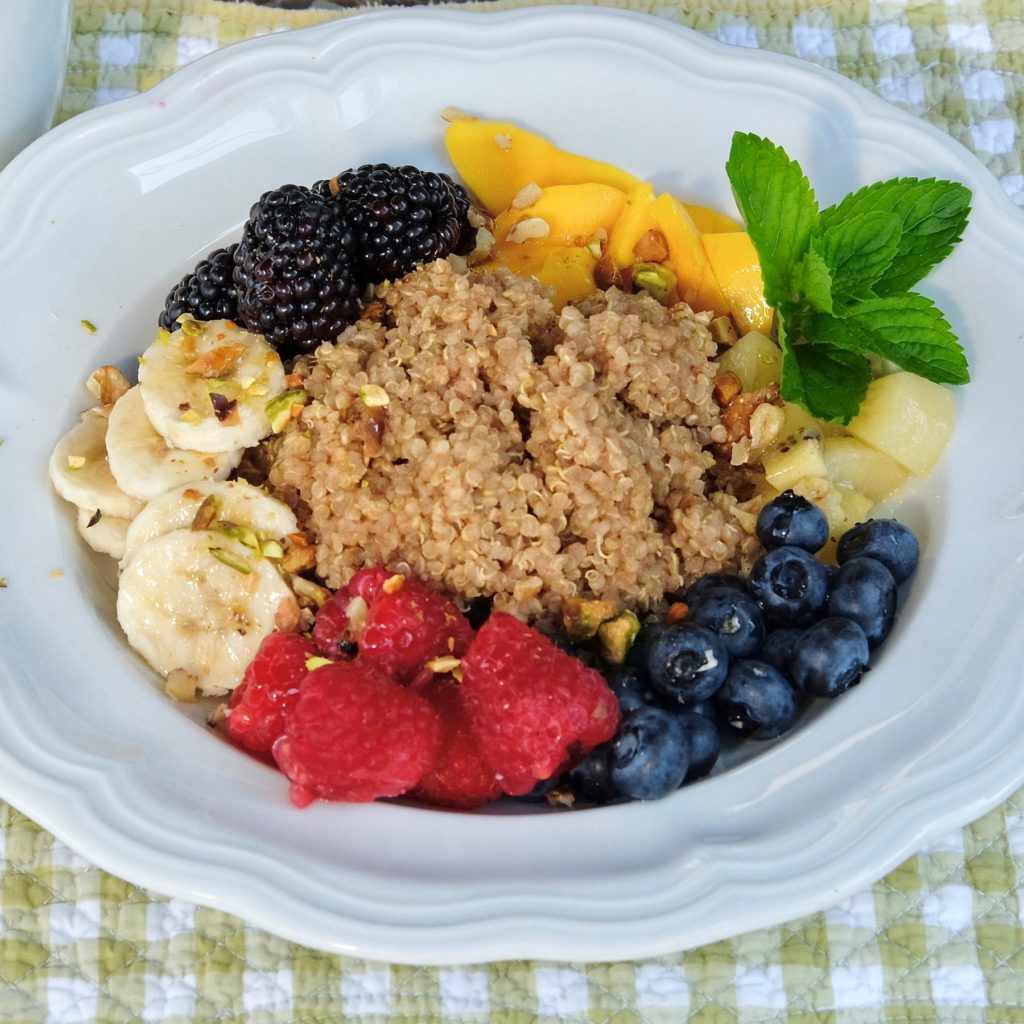 Quinoa in white bowl surrounded by blueberries, raspberries, sliced banana, blackberries and chopped mango, sprinkled with chopped pistachios