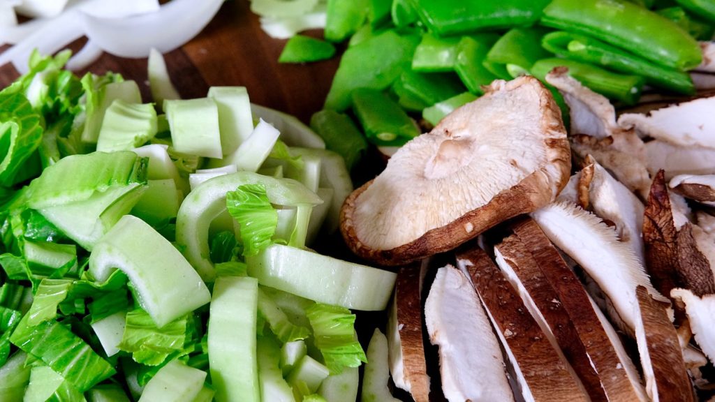 Chopped bok choy, sliced shitake mushroom and cleaned snap peas on walnut board.