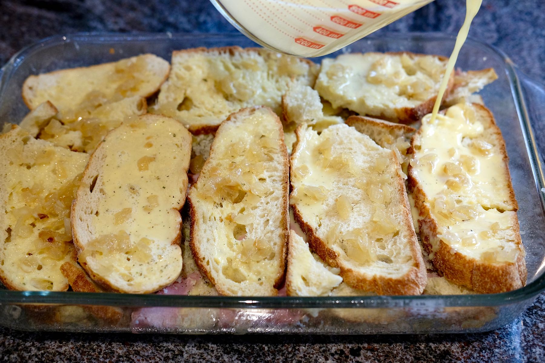 Layers of bread, ham and cheese  in glass baking dish while custard is being poured over.