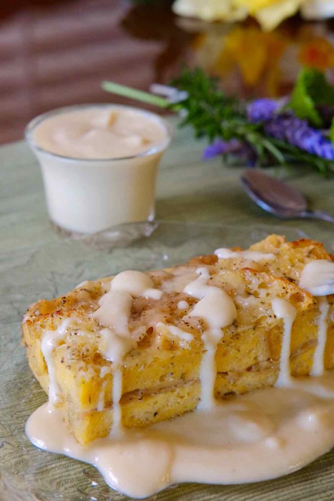 Ham and Gruyere Baked French Toast with Creamy Dijon Sauce on a clear glass plate with bowl of sauce and bouquet of lavenders in background. 