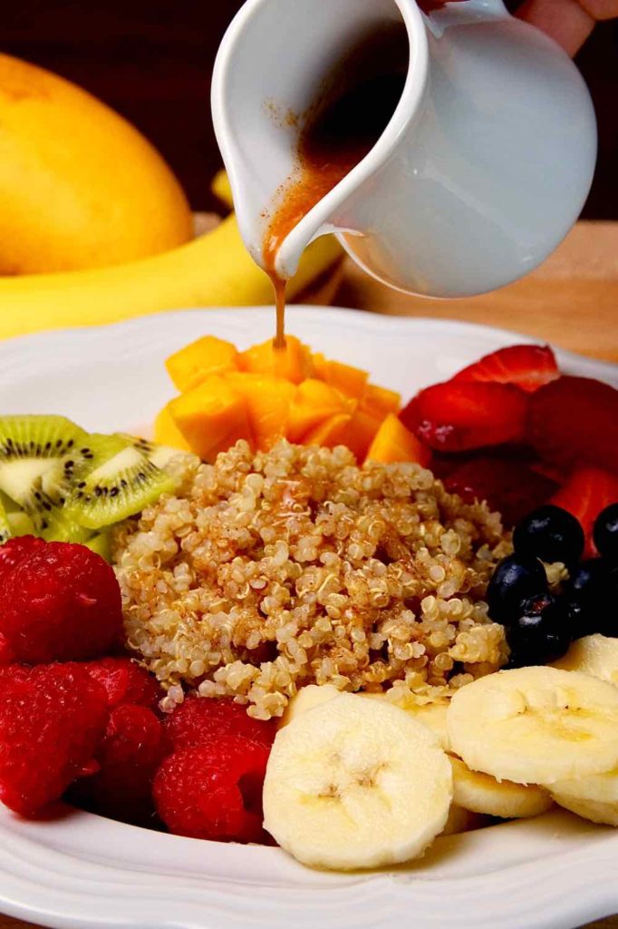 Quinoa Breakfast Bowl in a white bowl.  Quinoa drizzled with cinnamon-orange sauce being drizzled over the top and served with bananas, blueberries, strawberries, mango, kiwi and raspberries. Fresh bananas, mangos and kiwi in background.