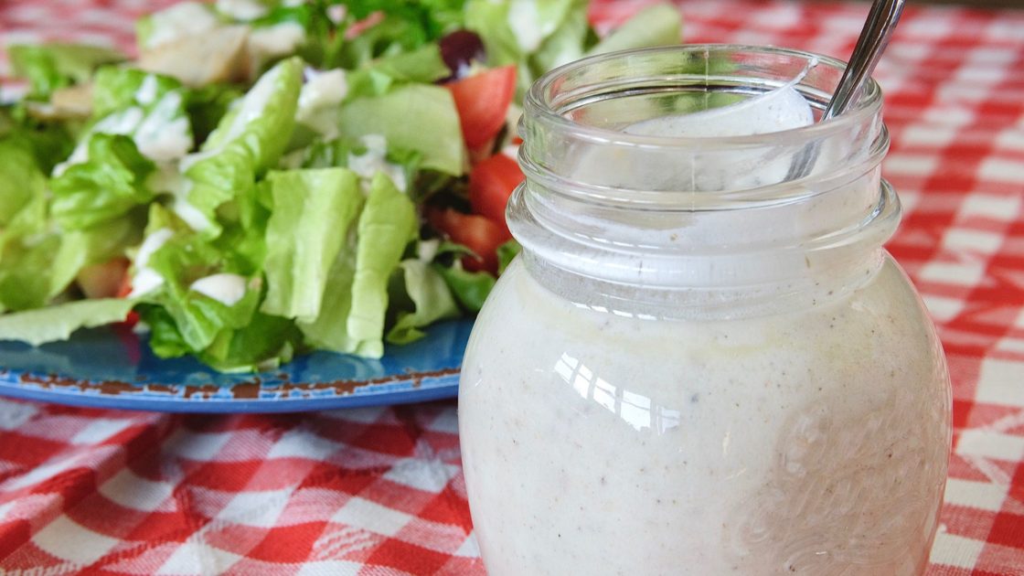 Creamy Greek Dressing in jar with salad on blue plate in background on a red and white checkered overlay