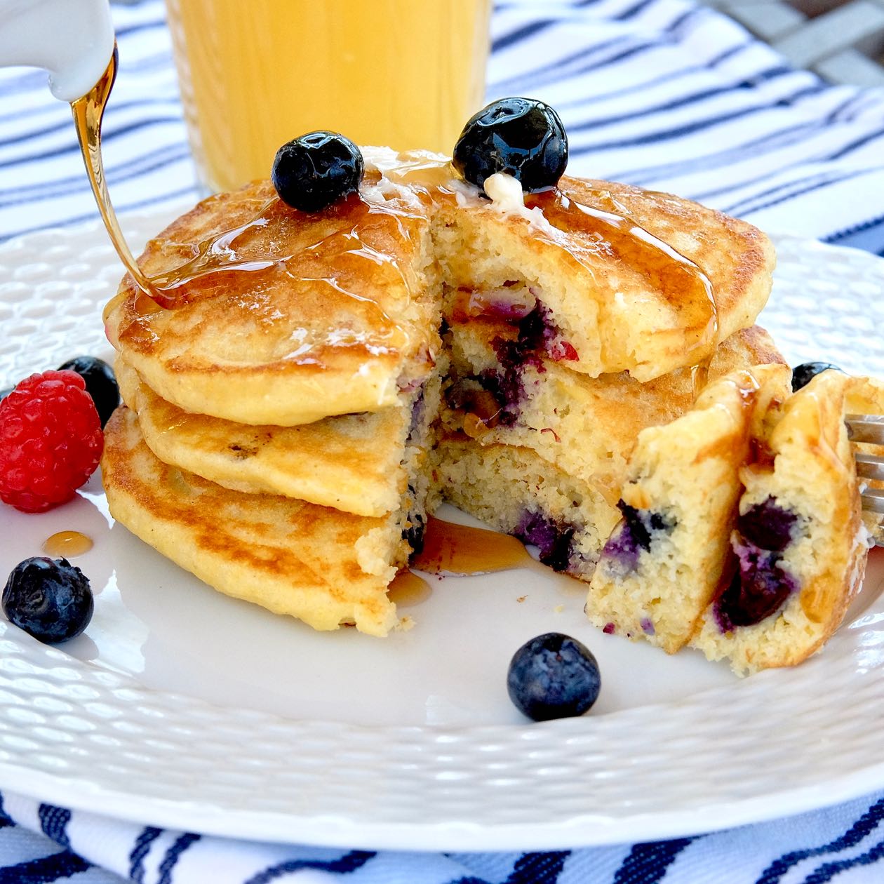 Blueberry Corn Cakes on white plate with syrup being poured over them. Glass of orange juice in background