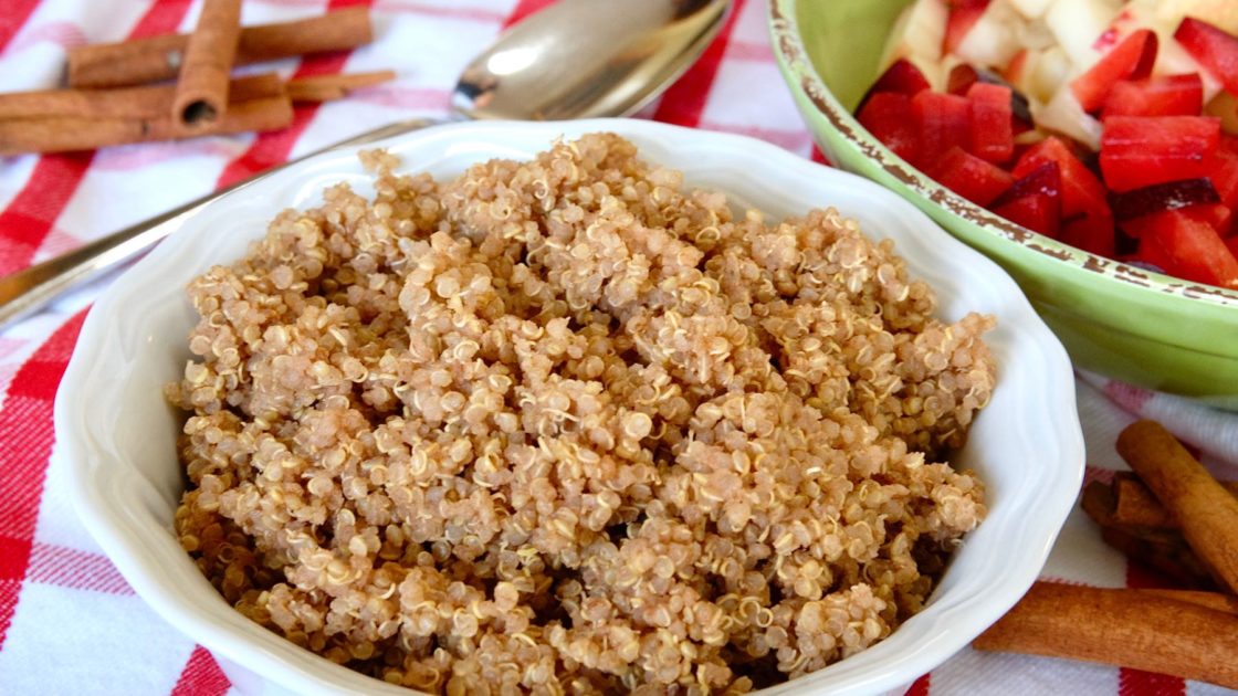 Cinnamon Almond Quinoa in white bowl and green bowl of chopped fruit on a red and white print overlay with cinnamon sticks in foreground