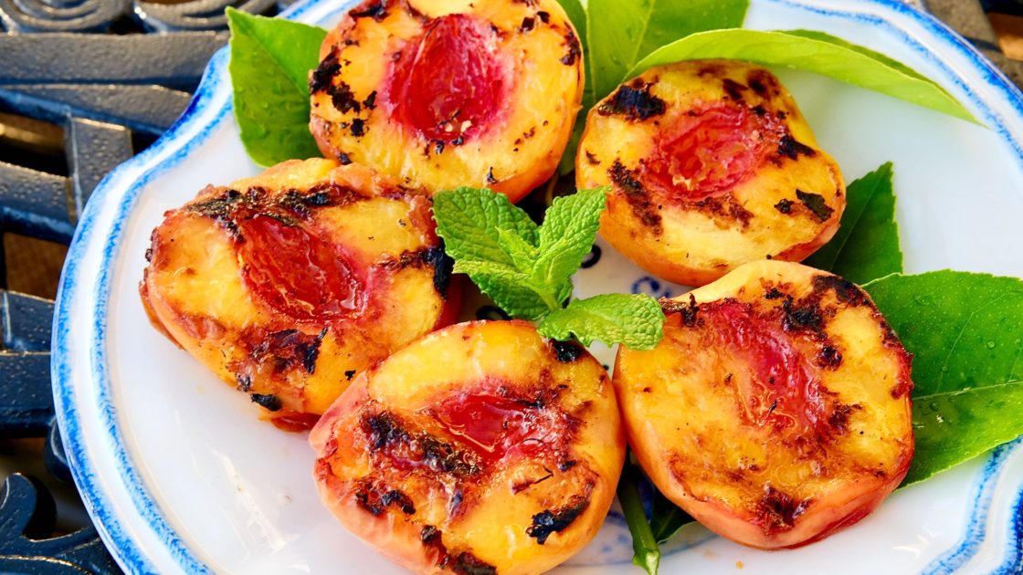 Grilled Peach halves displayed on a white plate with baby blue border. Lemon leaves make a colorful contrast displayed under the peaches.