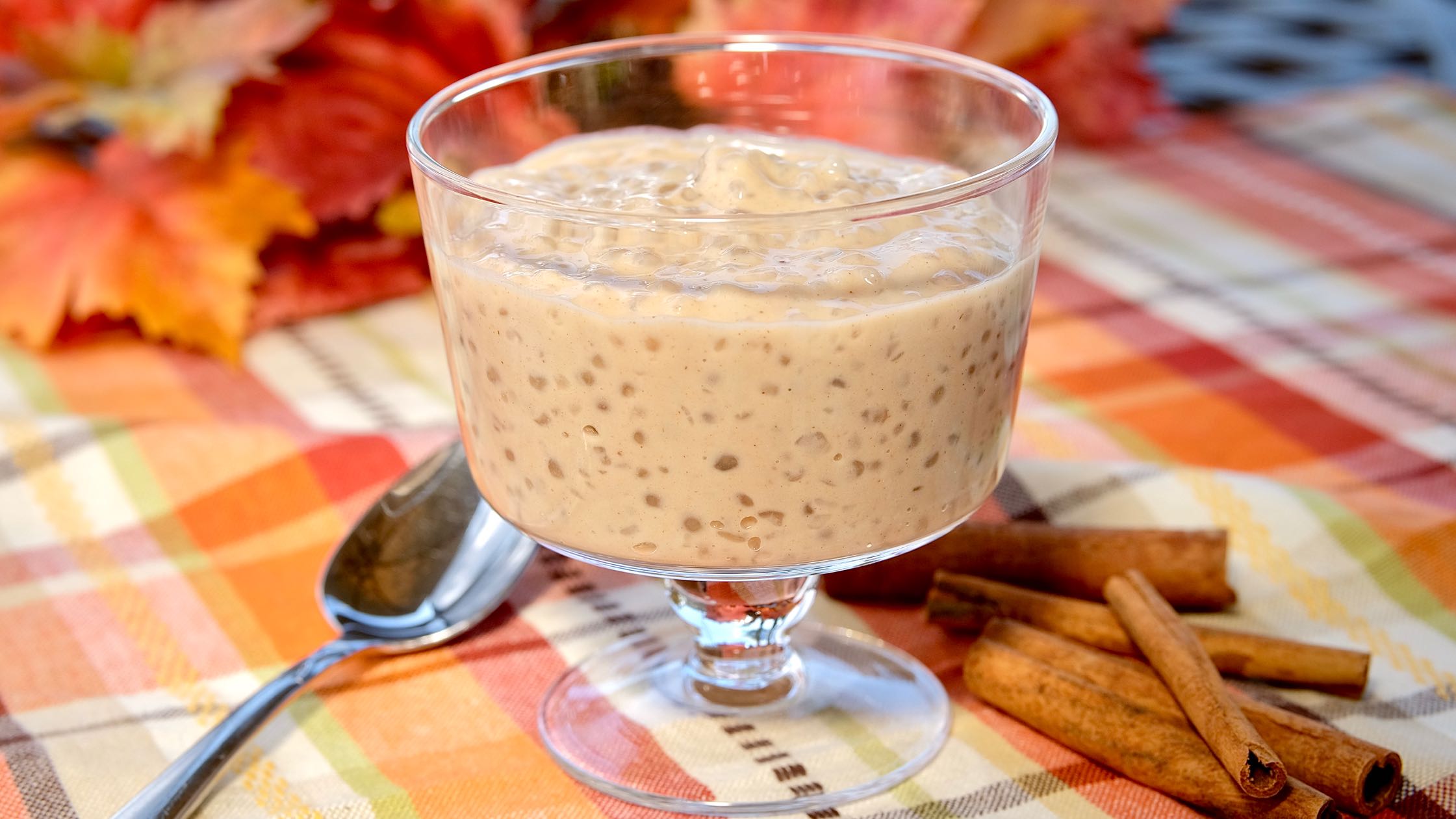 Cinnamon Tapioca Pudding in glass cup on overlay of orange and brown plaid with cinnamon sticks and spoon in foreground and autumn leave in background