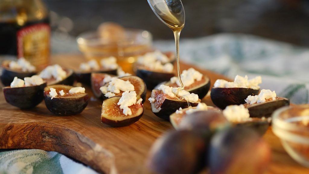 Fig appetizers served on an olive wood board with action shot of honey being drizzled.