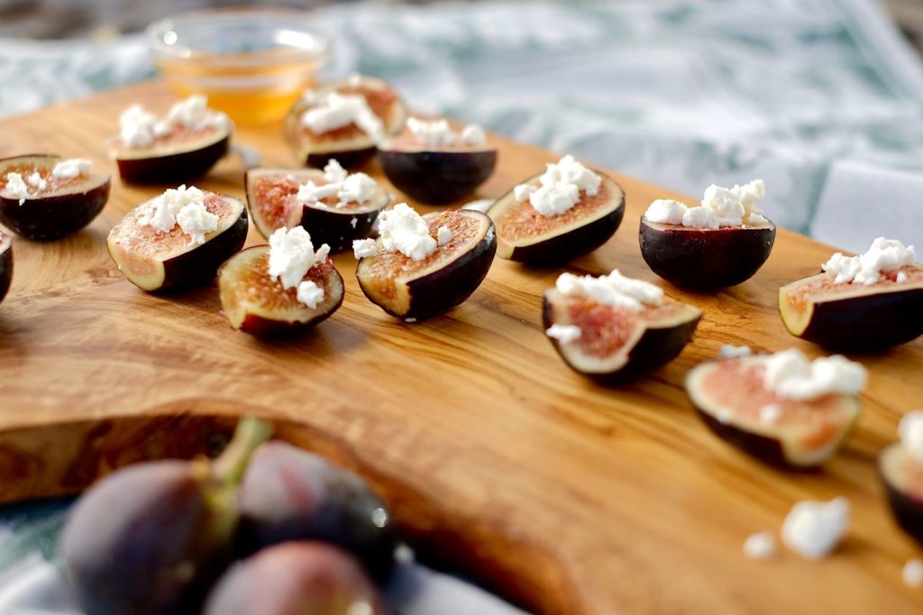 Fig halves dotted with crumbled goat cheese on Olive Board. A cluster of figs is in foreground.