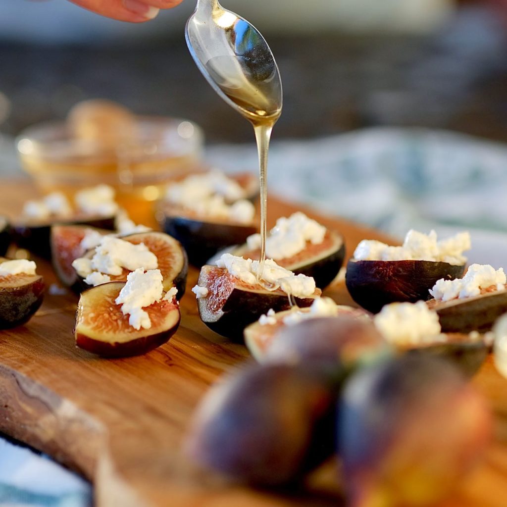 Figs with crumbled goat cheese are being drizzled with honey and served on an olive wood board.  