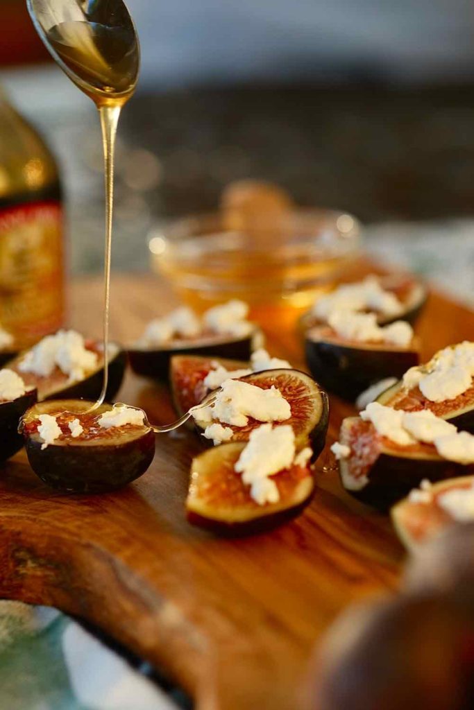 Easy Fig Appetizer topped with crumbled goat cheese and drizzled with honey served on an olivewood board with honey and wine bottle  in background.