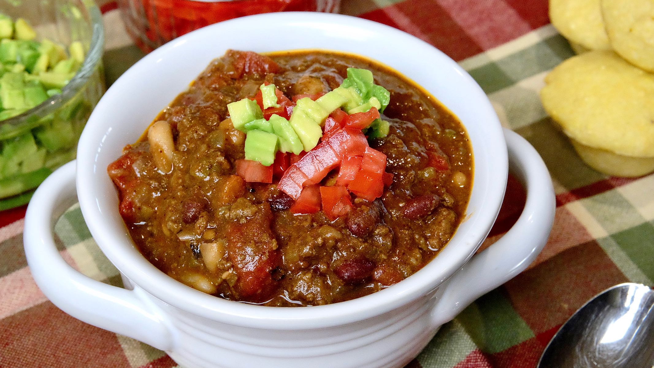 Hearty Bison Chocolate Chili in white bowl with chopped avocado and chopped tomato in background with corn muffins.