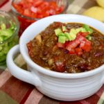 Hearty Bison Chocolate Chili in white chili bowl with chopped tomato and avocado in background.