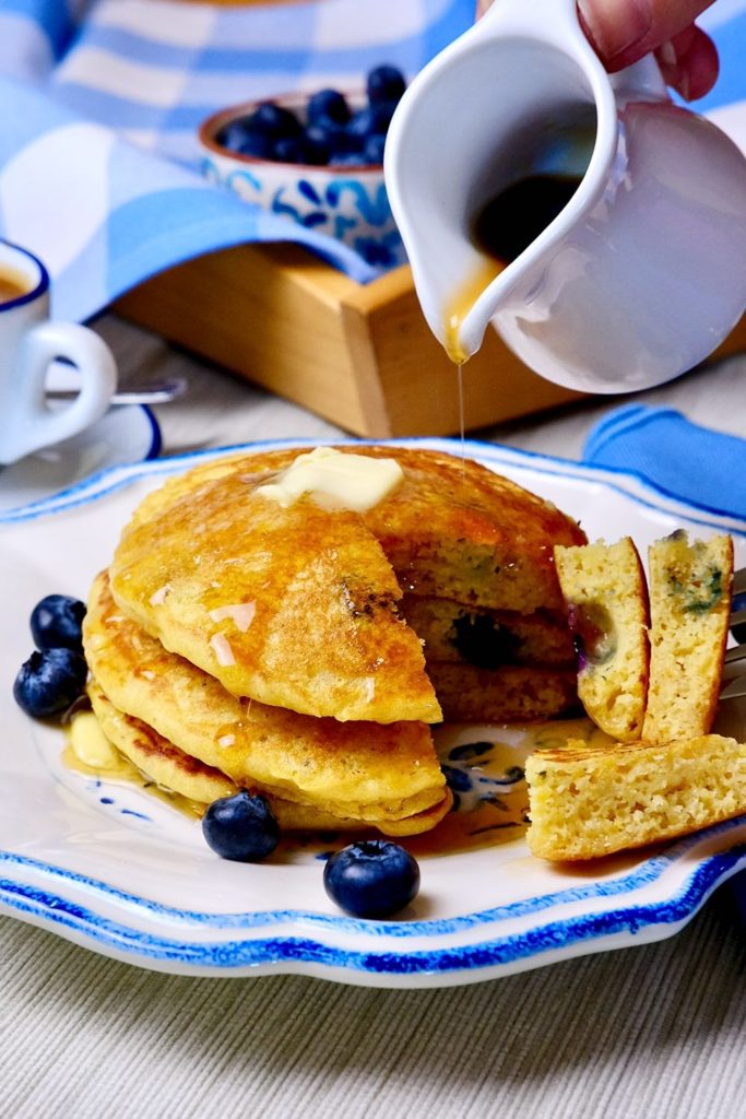 Sweet Corn Cakes on blue and white dish garnished with blueberries showing bite shot.  Plate is  set on beige linen with serving tray with blue and white linen and cup of espresso in background.   