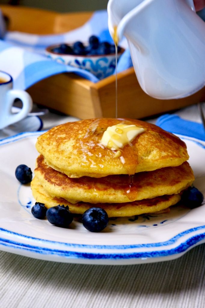 Sweet Corn Cakes on blue and white dish garnished with blueberries set on beige linen.  Serving tray with blue and white linen and cup of espresso in background>