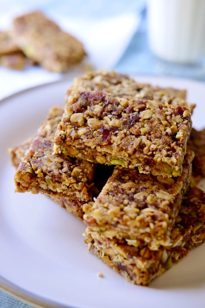 Pistachio-Apricot Granola bars stacked on white plate set on blue linen. Snack bar on napkin and glass of milk in background.