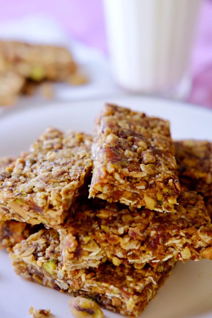 Granola bars stacked on white plate set on pink linen with glass of milk in background.