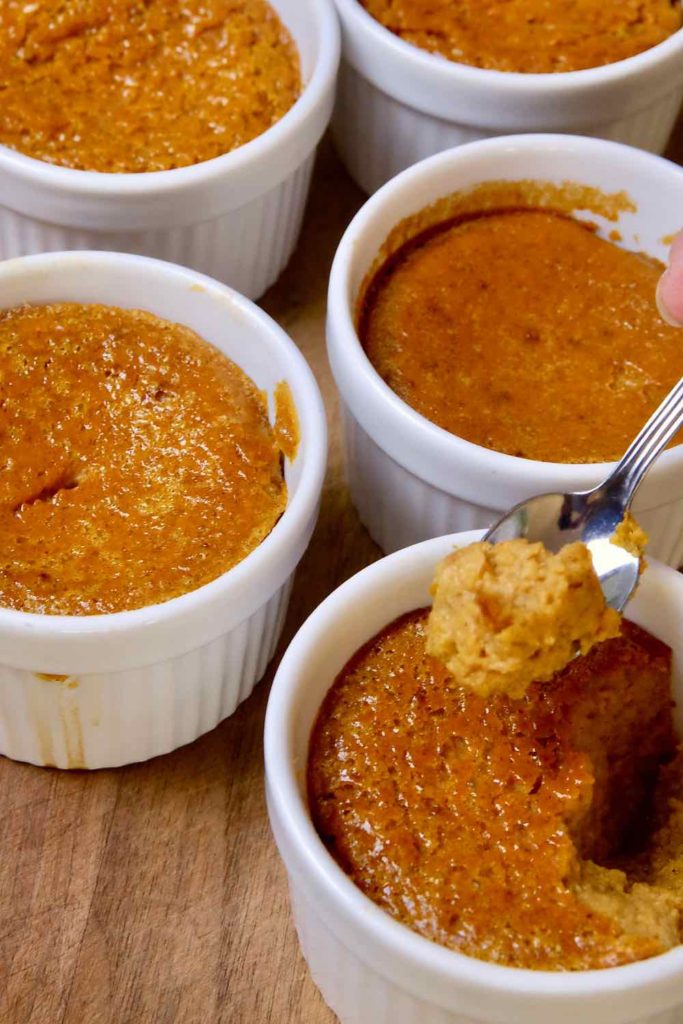 Pumpkin Custard baked in white ramekins set on walnut board.  One cup showing spoonful of a bite shot of custard. 