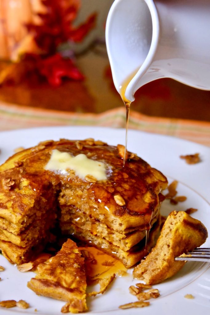 Fluffy Pumpkin Pancakes garnished with granola on white plate set on orange plaid linen.  Syrup is being poured over pancake bite shot.