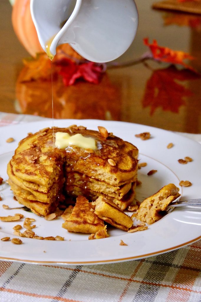 Fluffy Pumpkin Pancakes garnished with granola and drizzled with maple syrup served on ivory plate set on a fall harvest plaid linen.  Fall leave and pumpkin are in background. 
