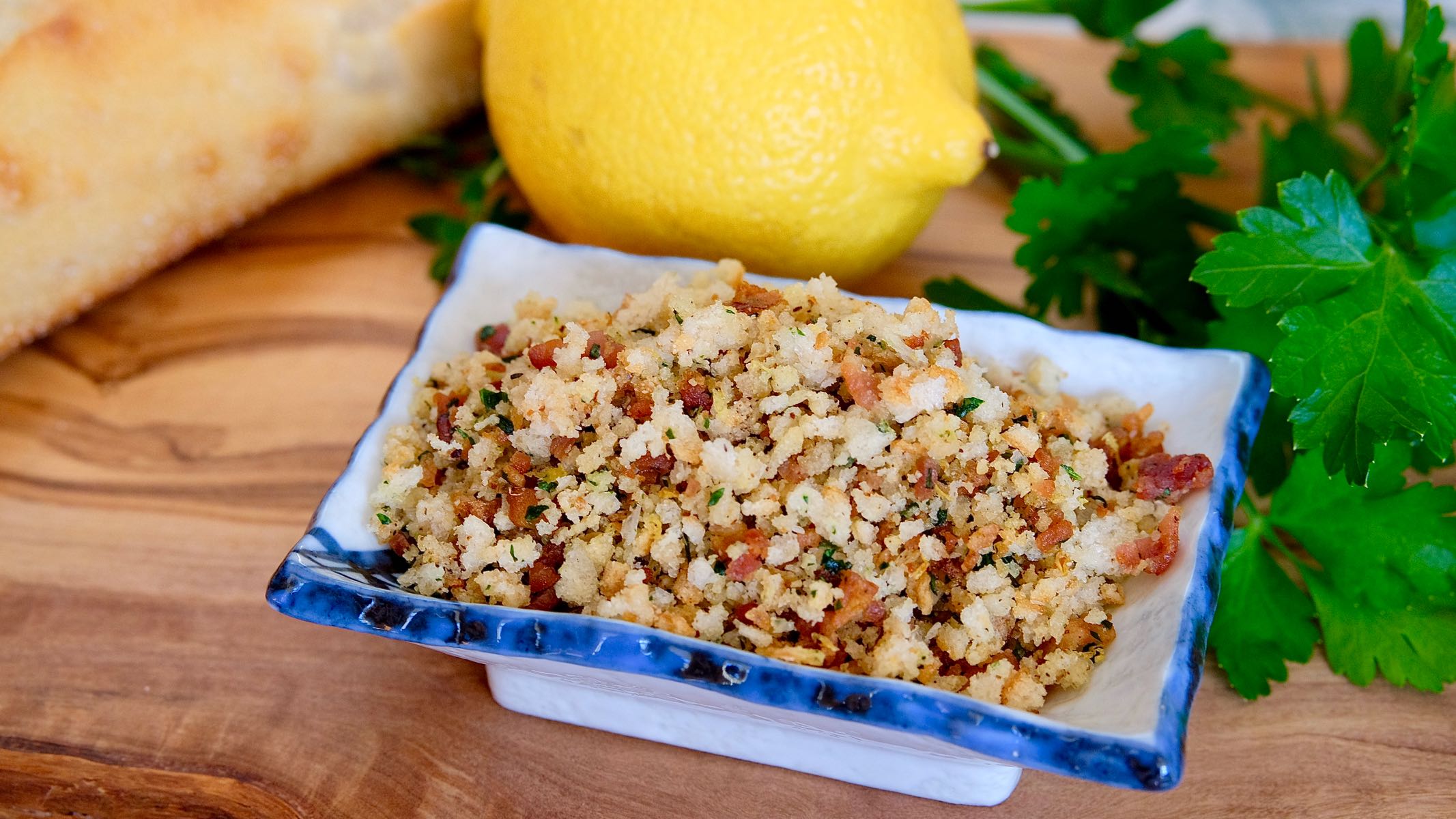 Toasted Breadcrumb Gremolata in square dish sitting on olive wood board with bread, lemon and parsley in background