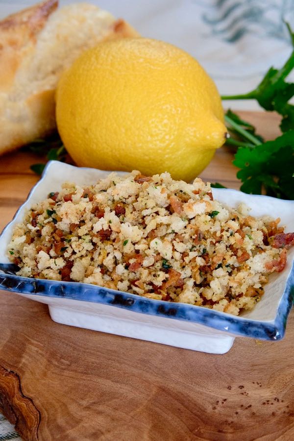 Toasted Breadcrumb Gremolata in square dish on olive wood board with bread, lemon and parsley in background
