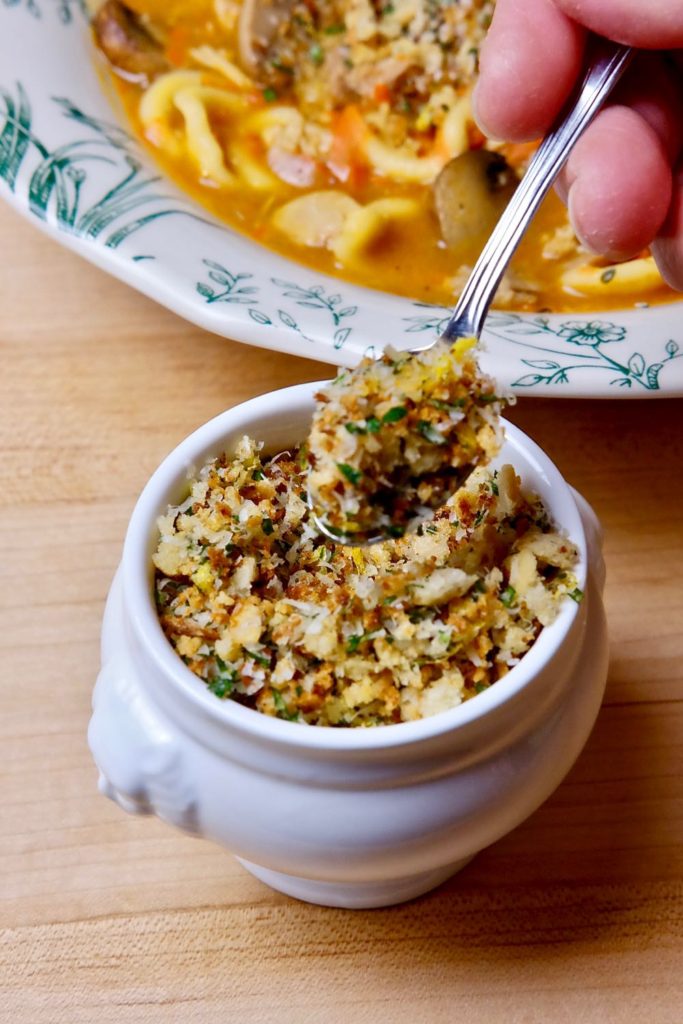 Toasted Breadcrumb gremolata in white bowl with spoon shot set on a maple board with bowl of soup in background.