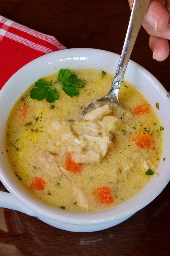 A spoonful of Greek-Style Lemon Chicken Soup in white two-handled soup bowl setting on dark wood underlay with red napkin in background