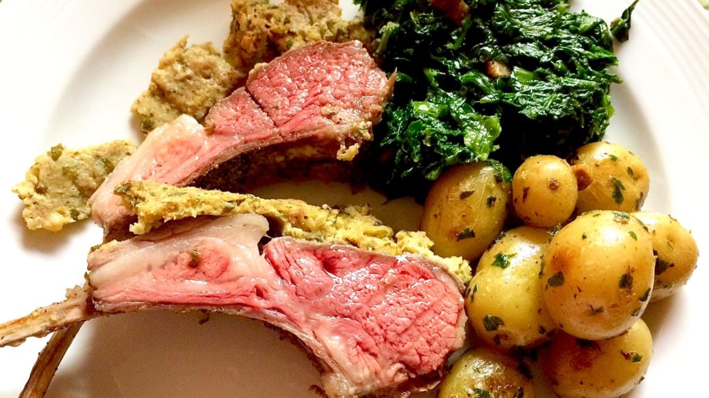 Two slices of rack of lamb set on white plate with sauteed spinach and parslied potatoes.