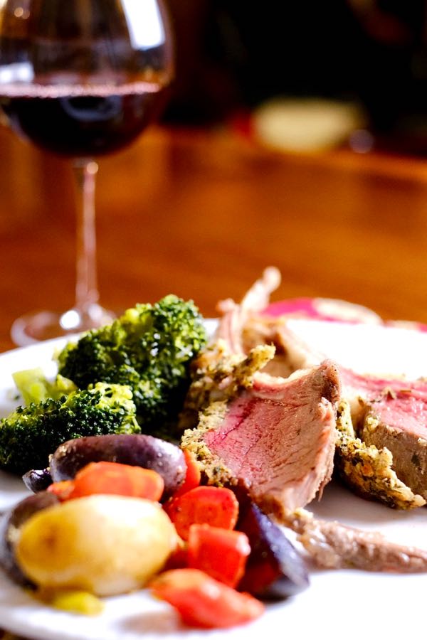 Herb-Crusted Rack of Lamb on white plate served with steamed broccoli, and Colorful Roasted Potatoes.  Glass of red wine in background.