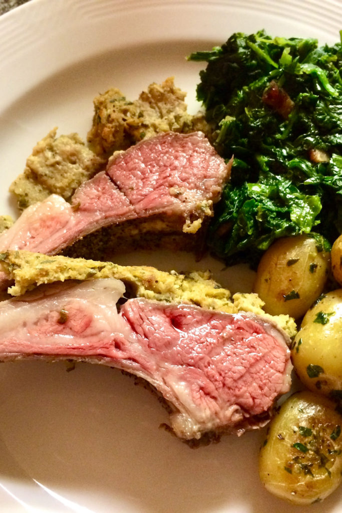 Two slices of herb crusted rack of lamb served on a white plate with parslied potatoes and sauteed spinach.