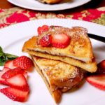 Nutella and Mascarpone Stuffed French Toast served with fresh sliced strawberries on a white plate with an additional plate of stacked french toast wedges in background.