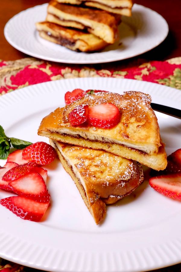 Nutella and Mascarpone Stuffed French Toast served with fresh sliced strawberries on a white plate with an additional plate of stacked french toast wedges in background.