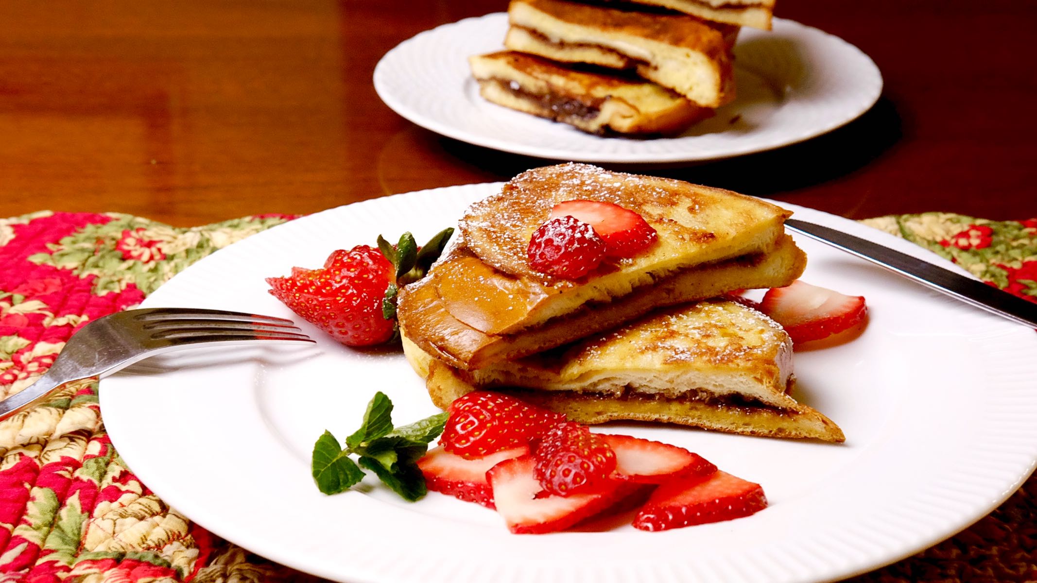 Nutella and Mascarpone Stuffed French toast on white plate garnished with sliced strawberries and stack of french toast in background