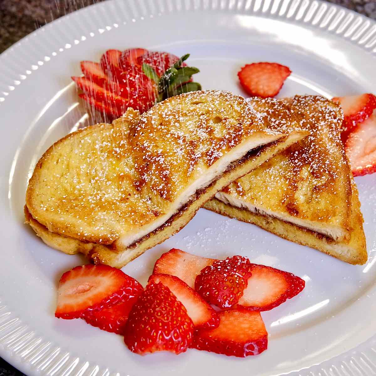 Nutella and Mascarpone Stuffed French Toast served with sliced strawberries on a white plate being dusted with confectioner's sugar.