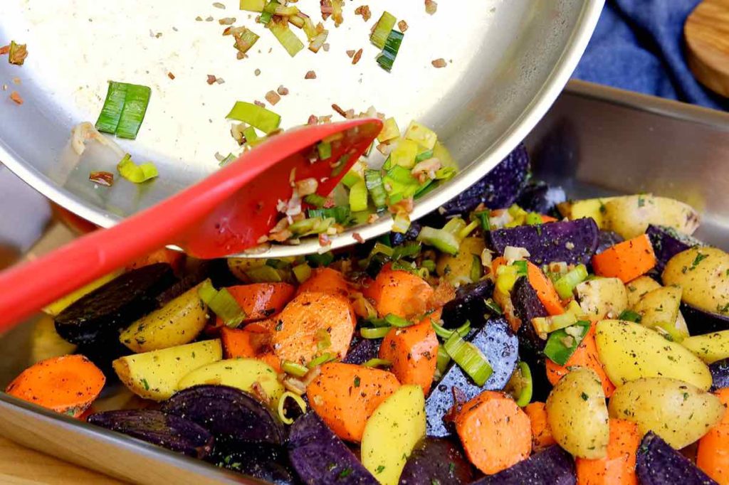 Sweet potatoes, purple and white baby potatoes and carrots being tossed with leek saute in baking pan.